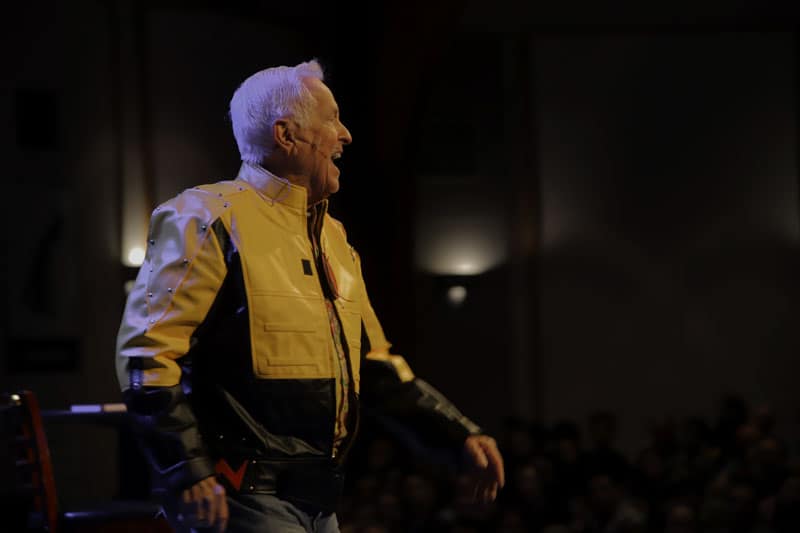 Josh McDowell, wearing a yellow jacket, speaking on stage at an apologetics event.