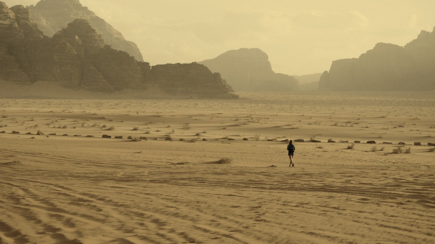 Person walking alone across a wide sandy desert under a cloudy sky